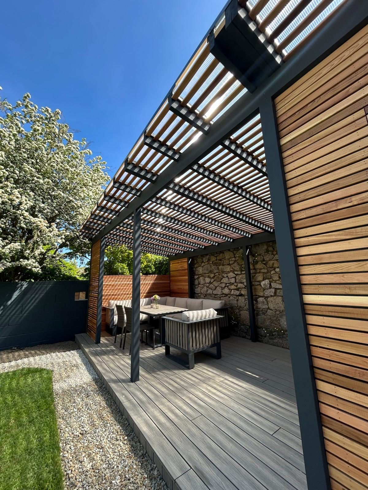 Pergola corner view — slatted cedar roof, stone wall and composite decking